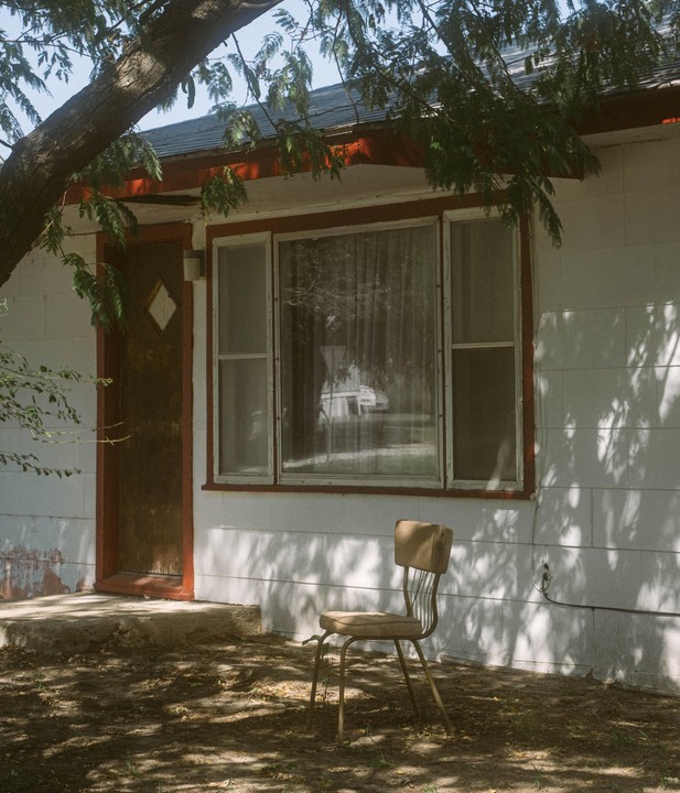 A single chair sits outside a white house with a red door frame and a large window, partially shaded by a tree.