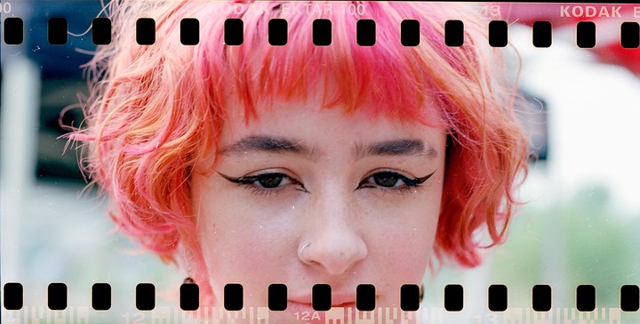 A woman with pink hair and winged eyeliner looks directly at the viewer.