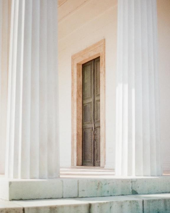 A pair of large white columns frame a dark door with intricate patterns, set within a light-colored building.
