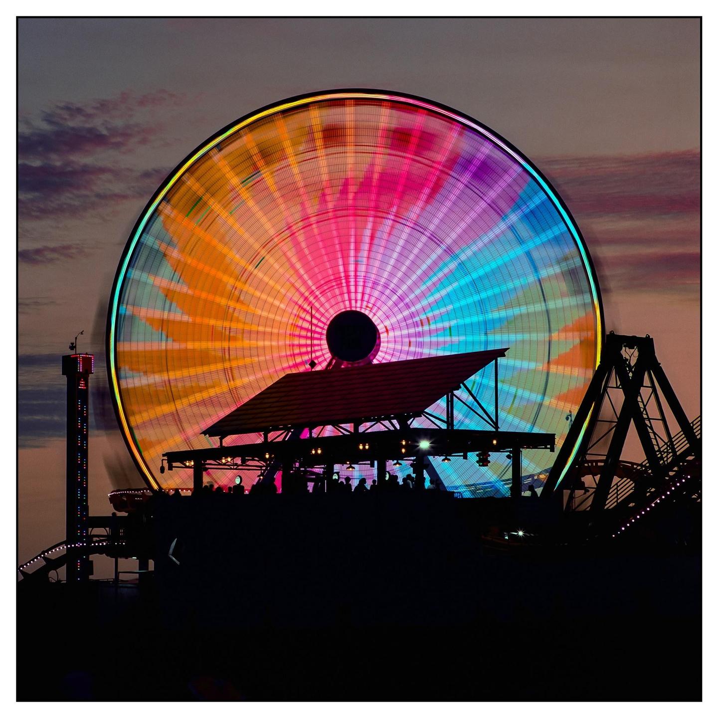 A colorful ferris wheel spins against a dusky sky above a silhouetted crowd and amusement park rides.