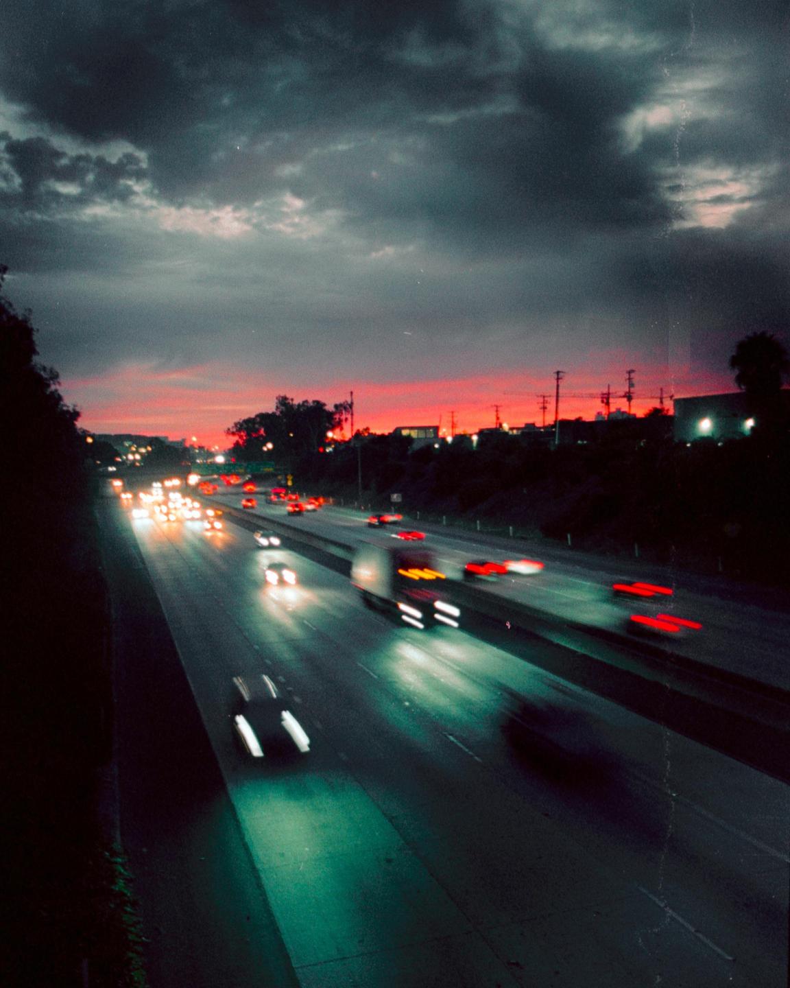 A highway at dusk shows streaks of light from moving vehicles under a dramatic sky.