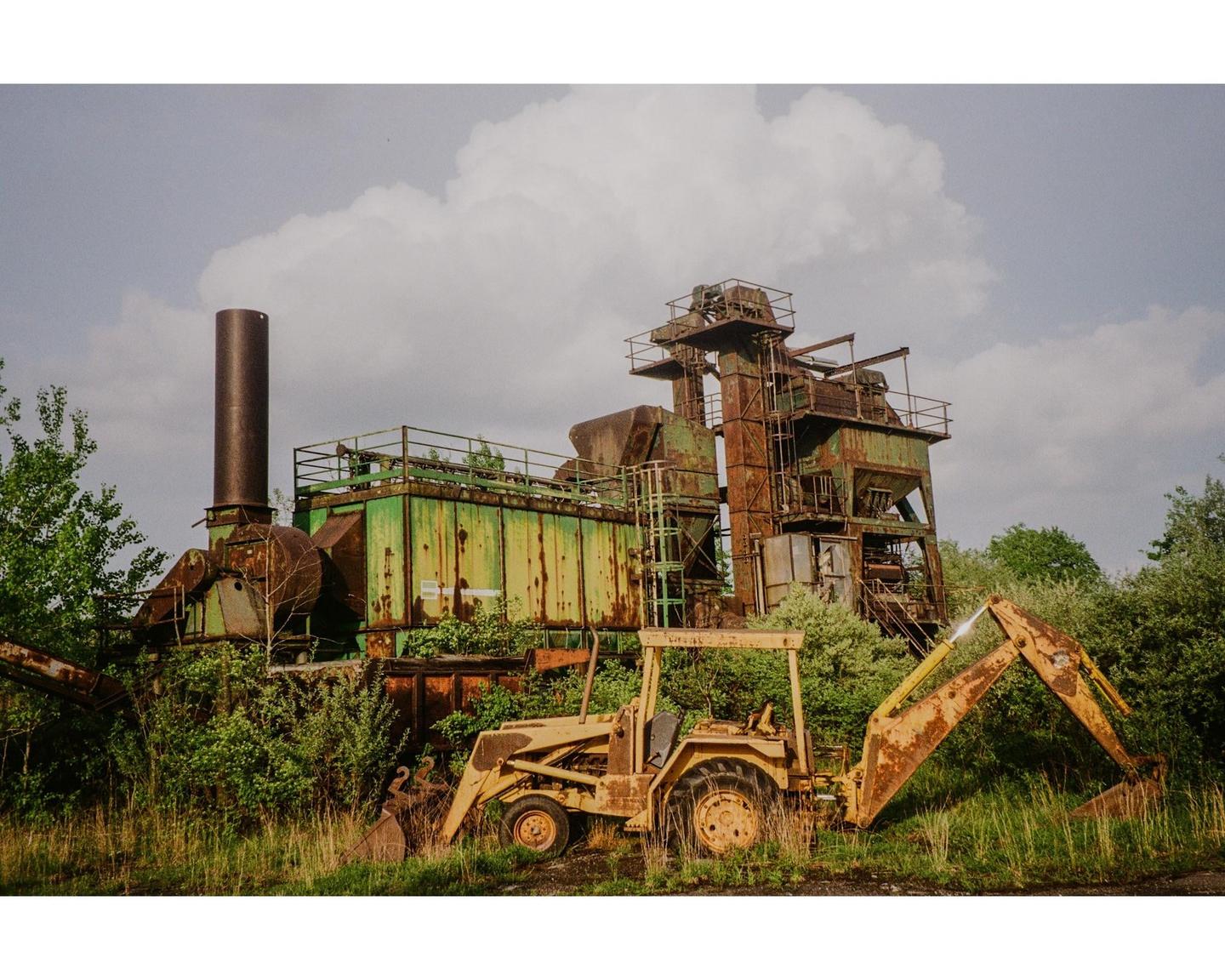 An abandoned industrial building and a rusty yellow excavator are overgrown with green foliage under a cloudy sky.