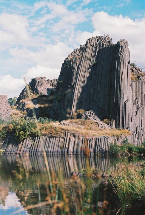 A large rock formation with vertical columns rises from the edge of a body of water under a blue sky with clouds.