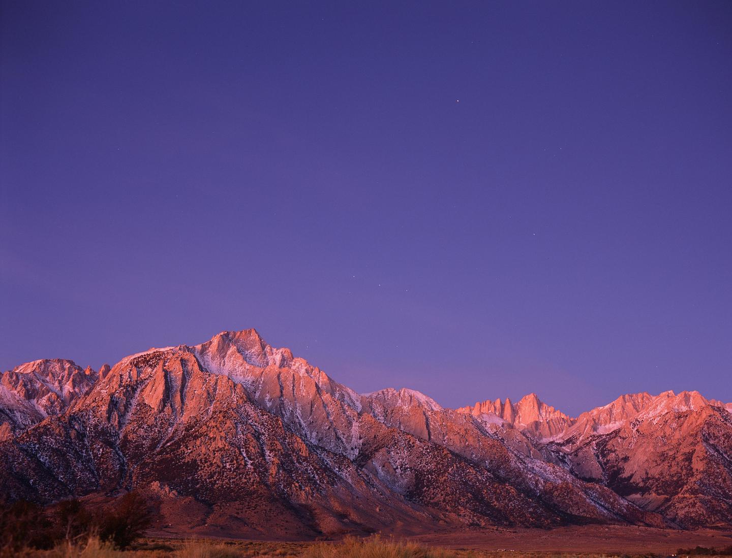 Snow-capped mountains are illuminated by the setting sun under a deep blue sky.