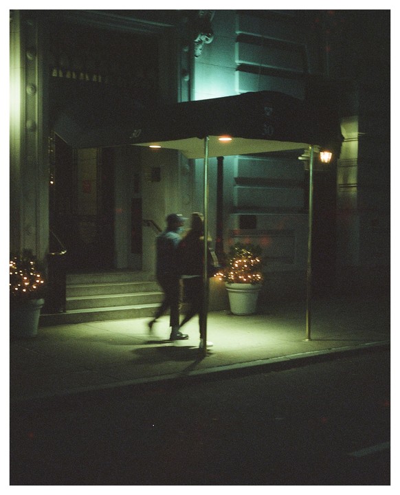 Two people walk past a building entrance with an awning and potted plants with lights at night.
