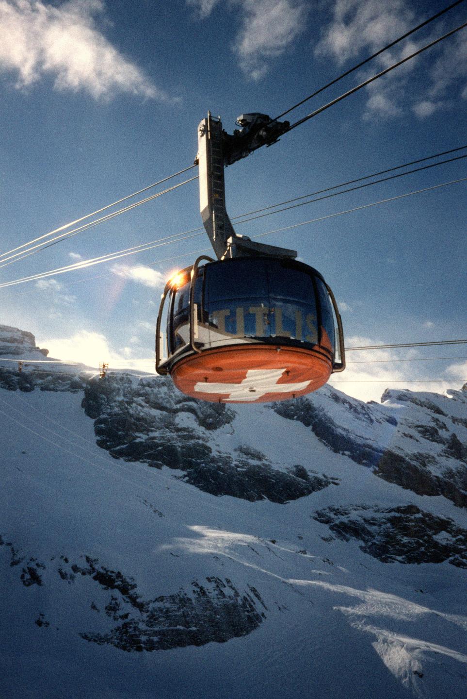 A cable car with a Swiss flag on its underside is suspended over a snow-covered mountain landscape under a blue sky with clouds.