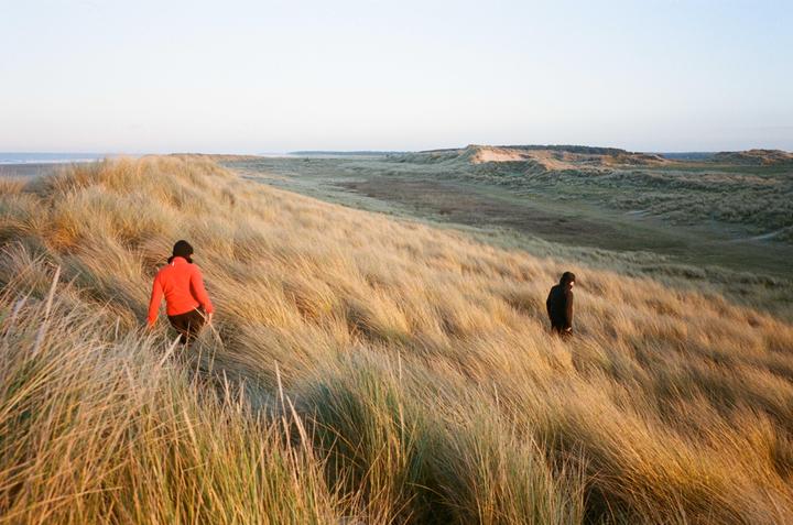Two people walk through tall grass on a dune with the ocean visible in the distance.