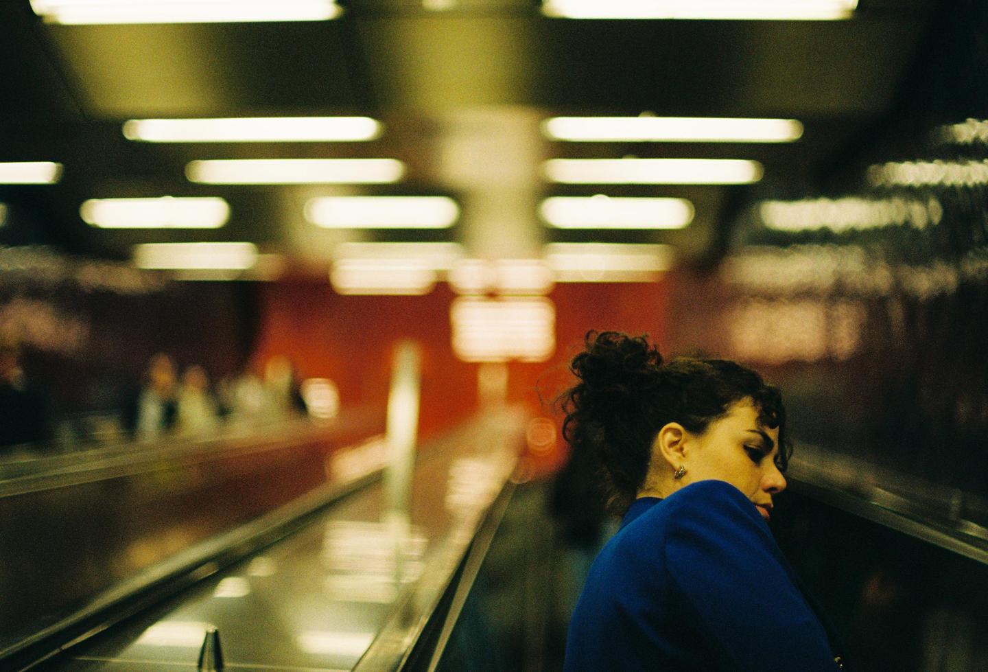 A woman with dark curly hair in a blue top stands on an escalator with blurred lights in the background.