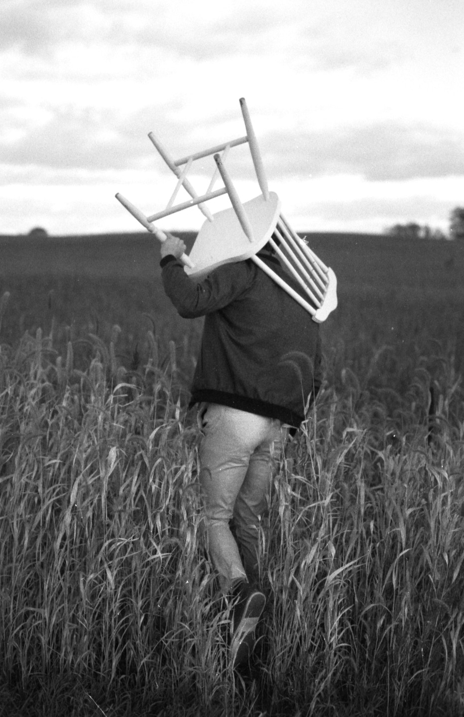 A man carries a chair on his shoulder through a field of tall grass.