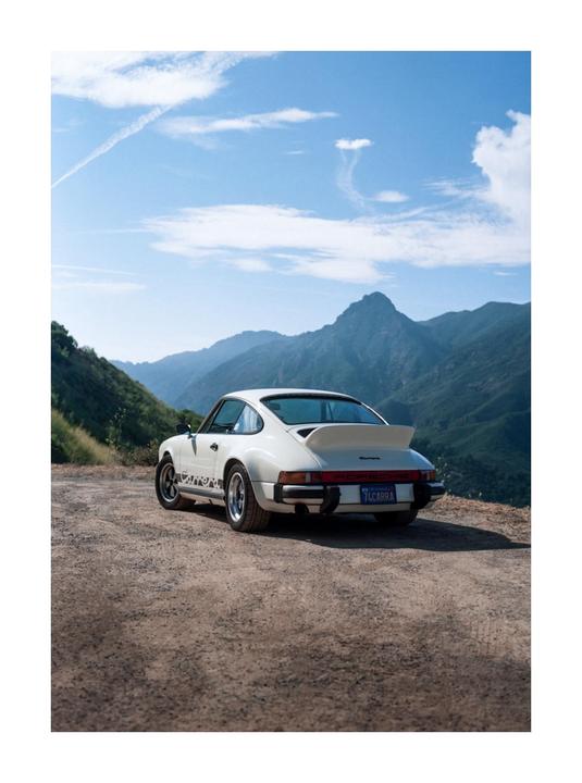 A white sports car is parked on a dirt road overlooking a mountain range under a blue sky.