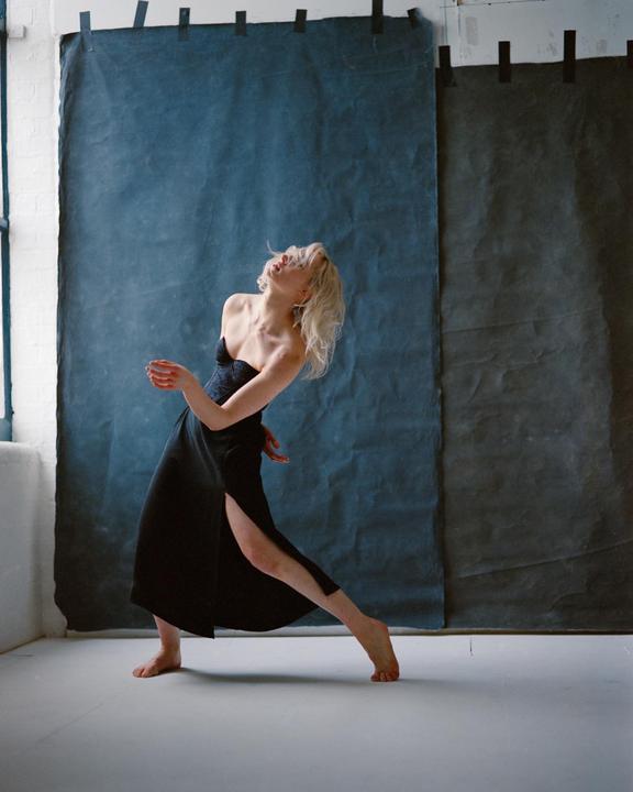 A woman with blonde hair and a black dress is dancing barefoot in a studio with a blue backdrop.