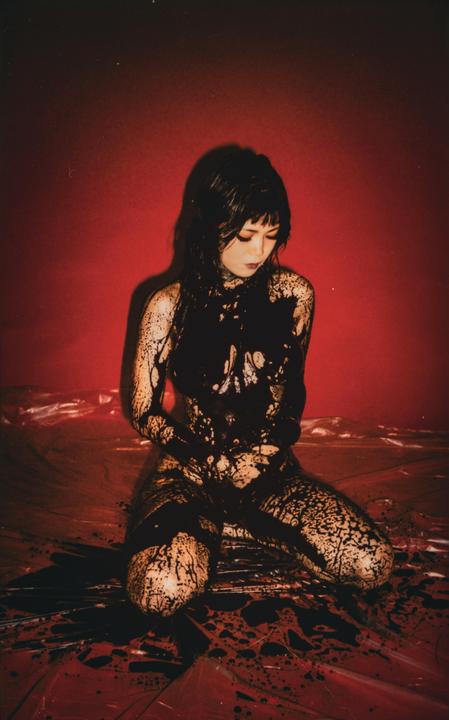 A woman kneels on a red surface, covered in black paint or liquid.