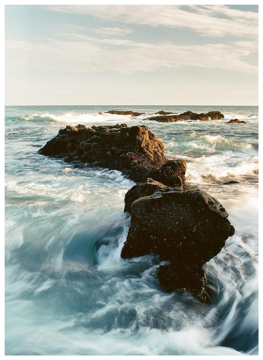 Waves crash around several large rocks in the ocean under a partly cloudy sky.