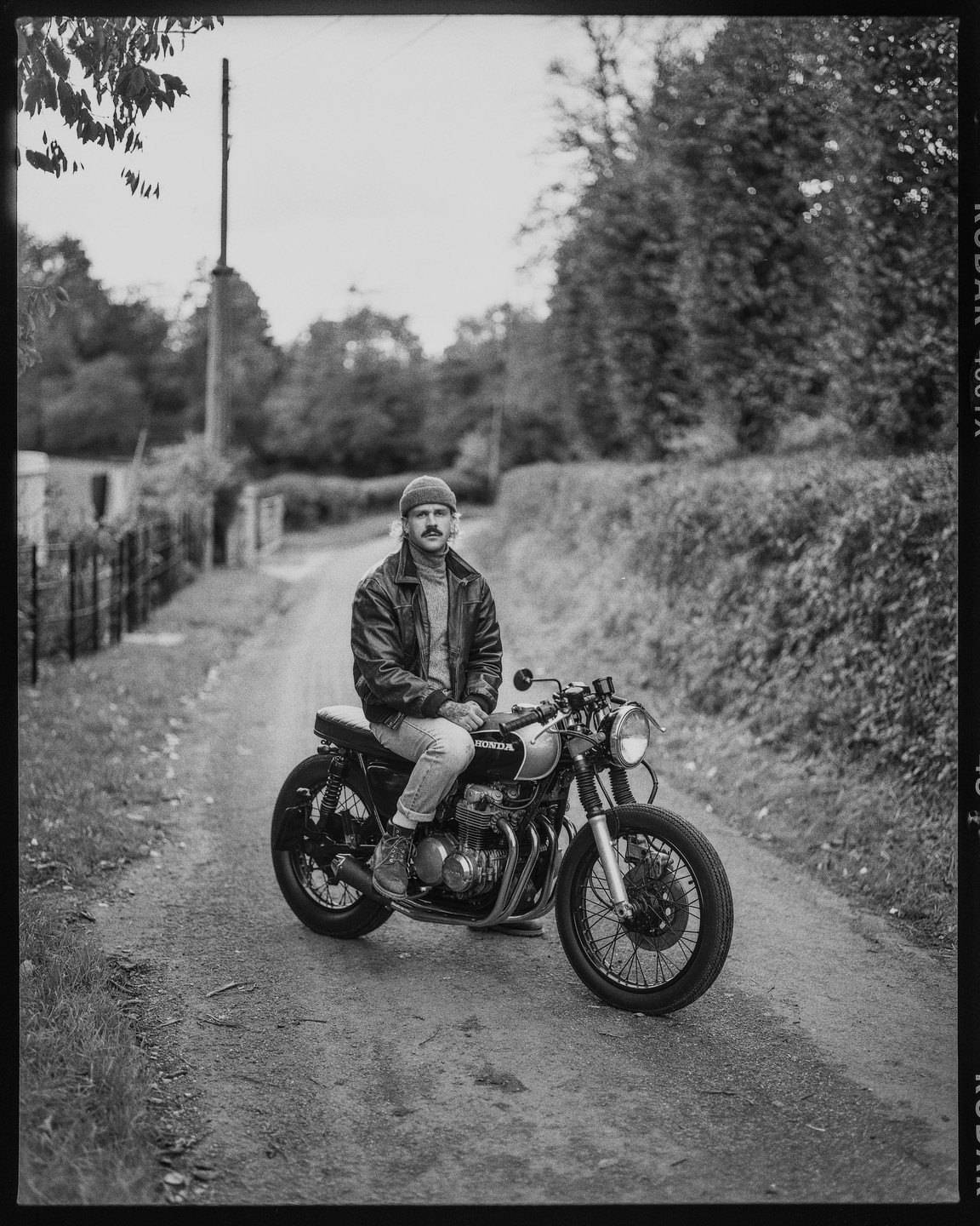 A man sits on a motorcycle on a dirt road lined with hedges and trees.