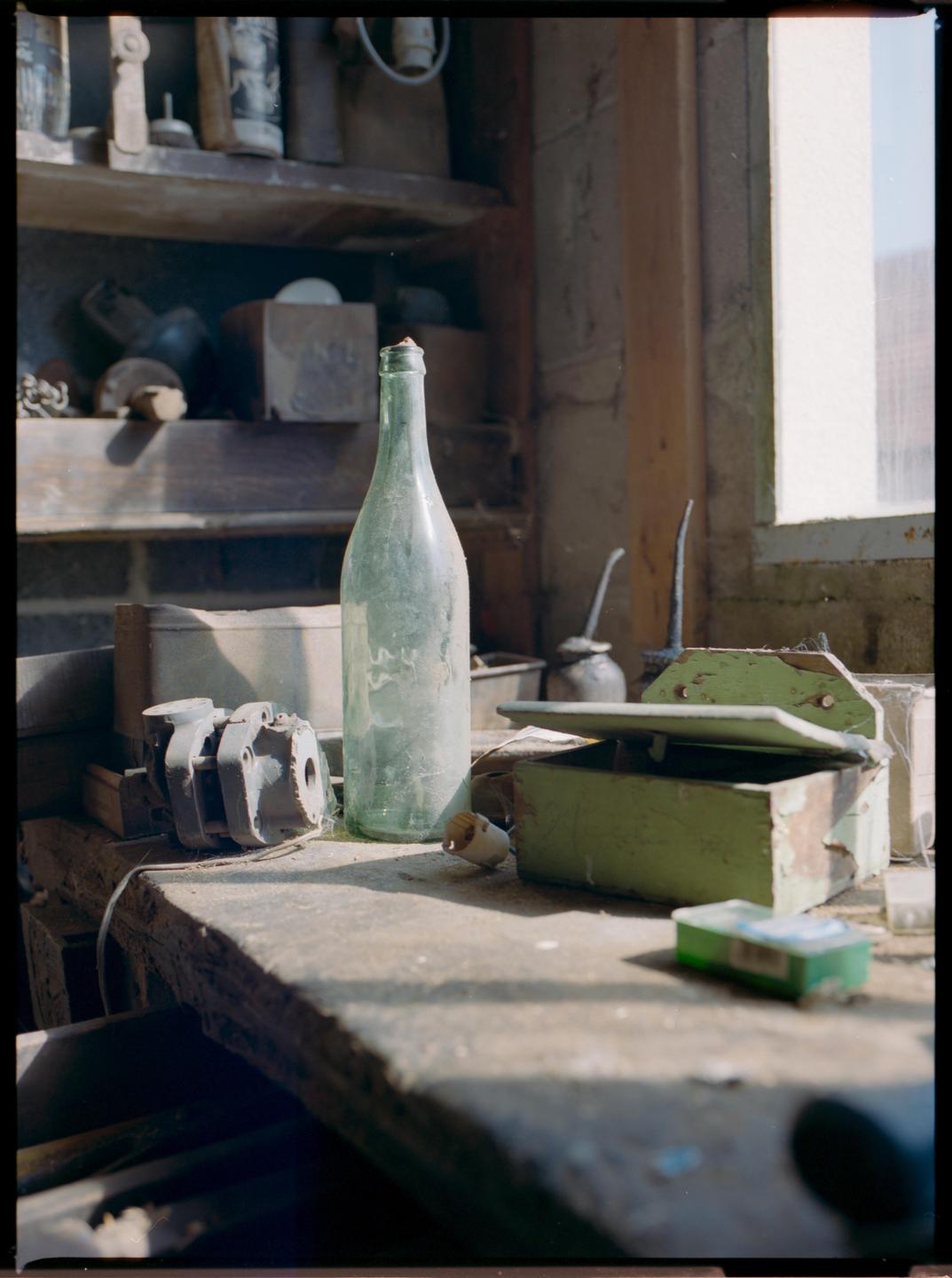 A dusty green bottle sits on a workbench next to an open green box and other assorted items.