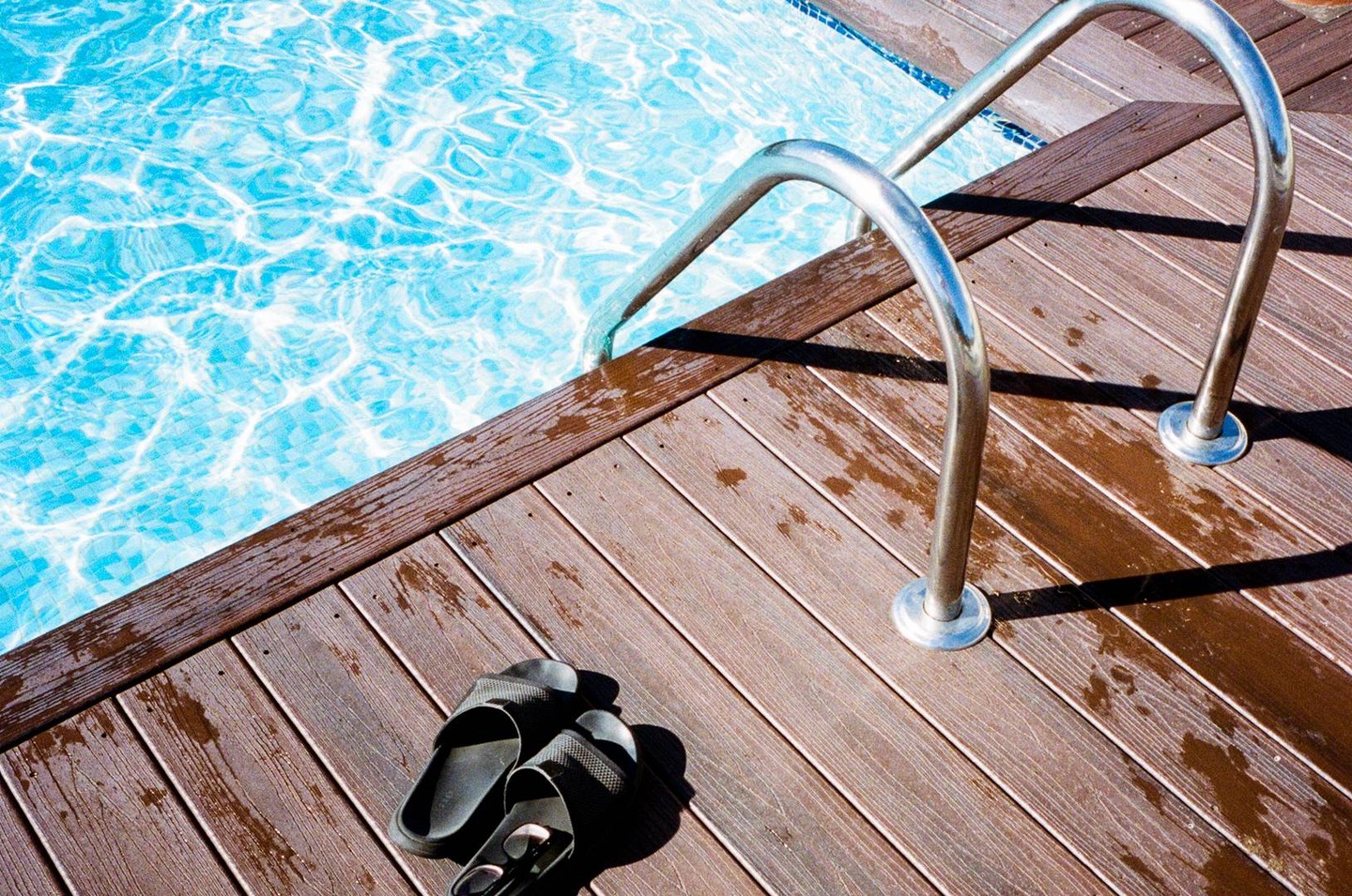 Black sandals rest on a wet wooden deck next to a swimming pool with a metal handrail.