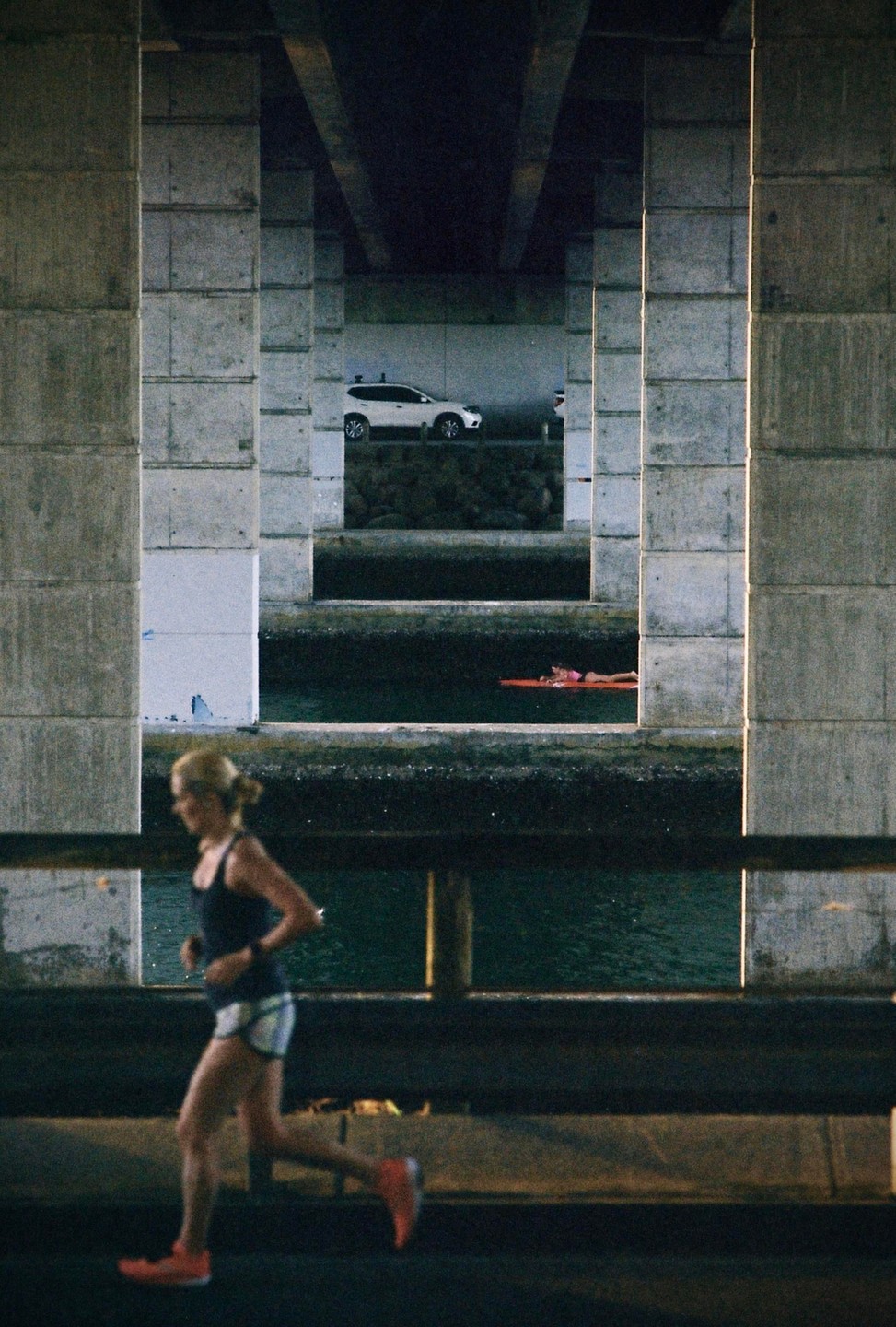 A woman runs along a road under a bridge, with a car and a person on a paddleboard visible in the distance.