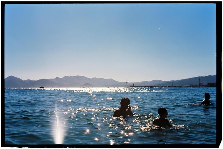 Three people are swimming in the sea with mountains and a pier in the background.