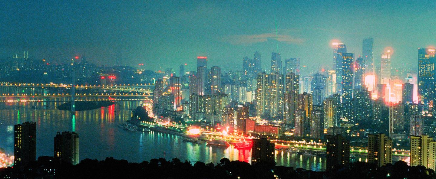 A panoramic night cityscape shows a river with bridges and illuminated buildings reflecting in the water.