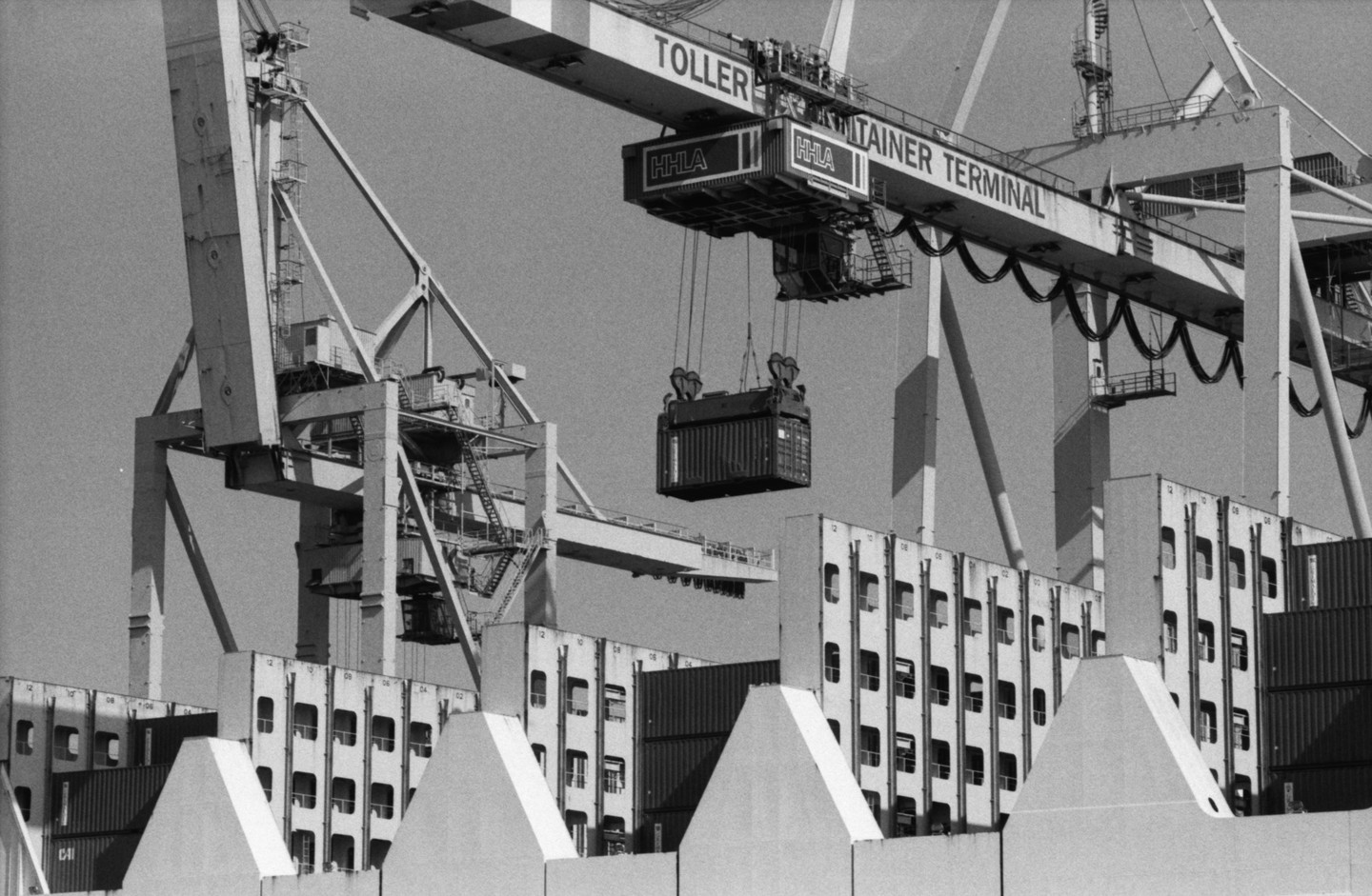 A large crane lifts a container over a ship filled with other containers at a terminal.