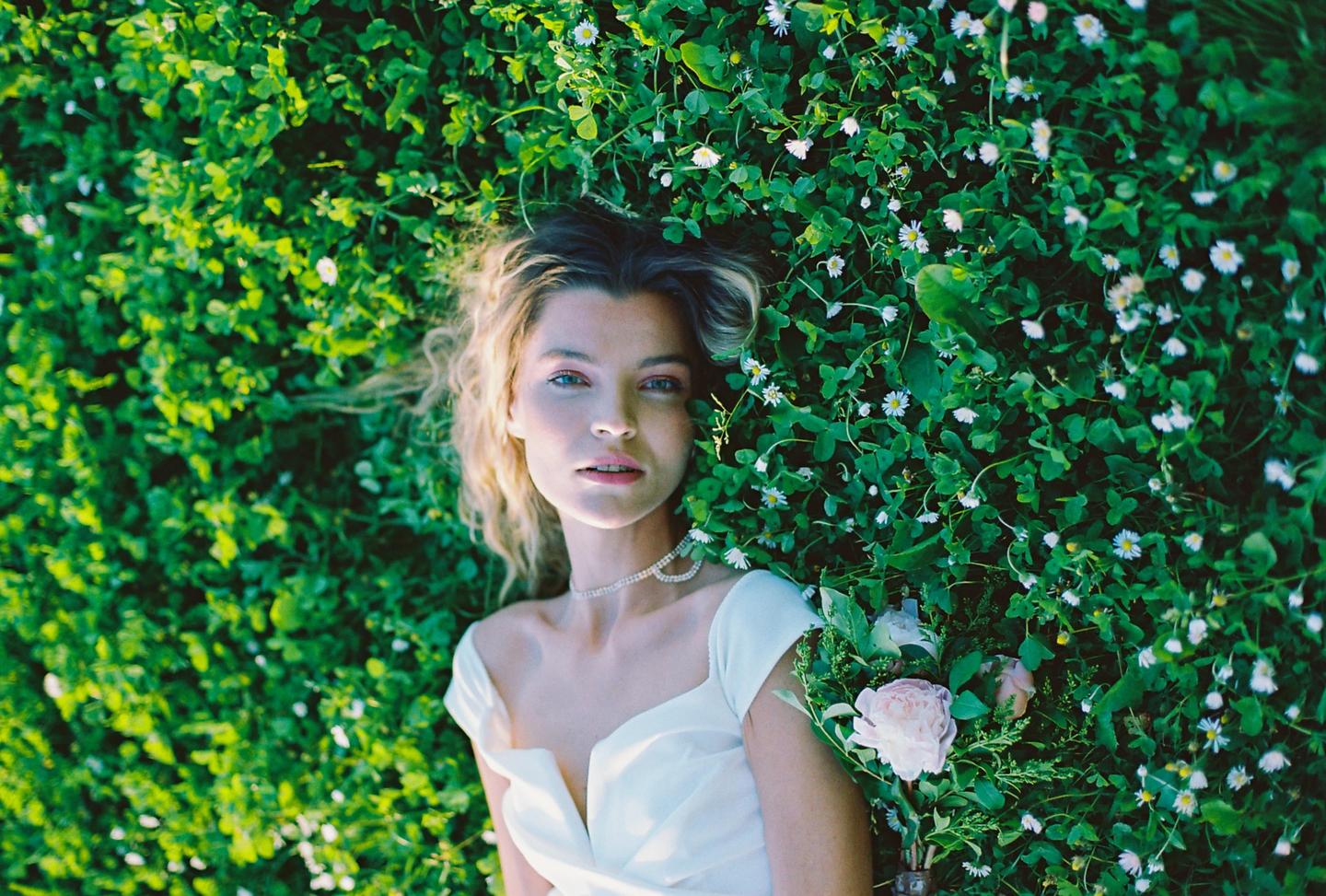 A woman in a white dress lies in a field of green clover and small white flowers, holding a bouquet of pink roses.