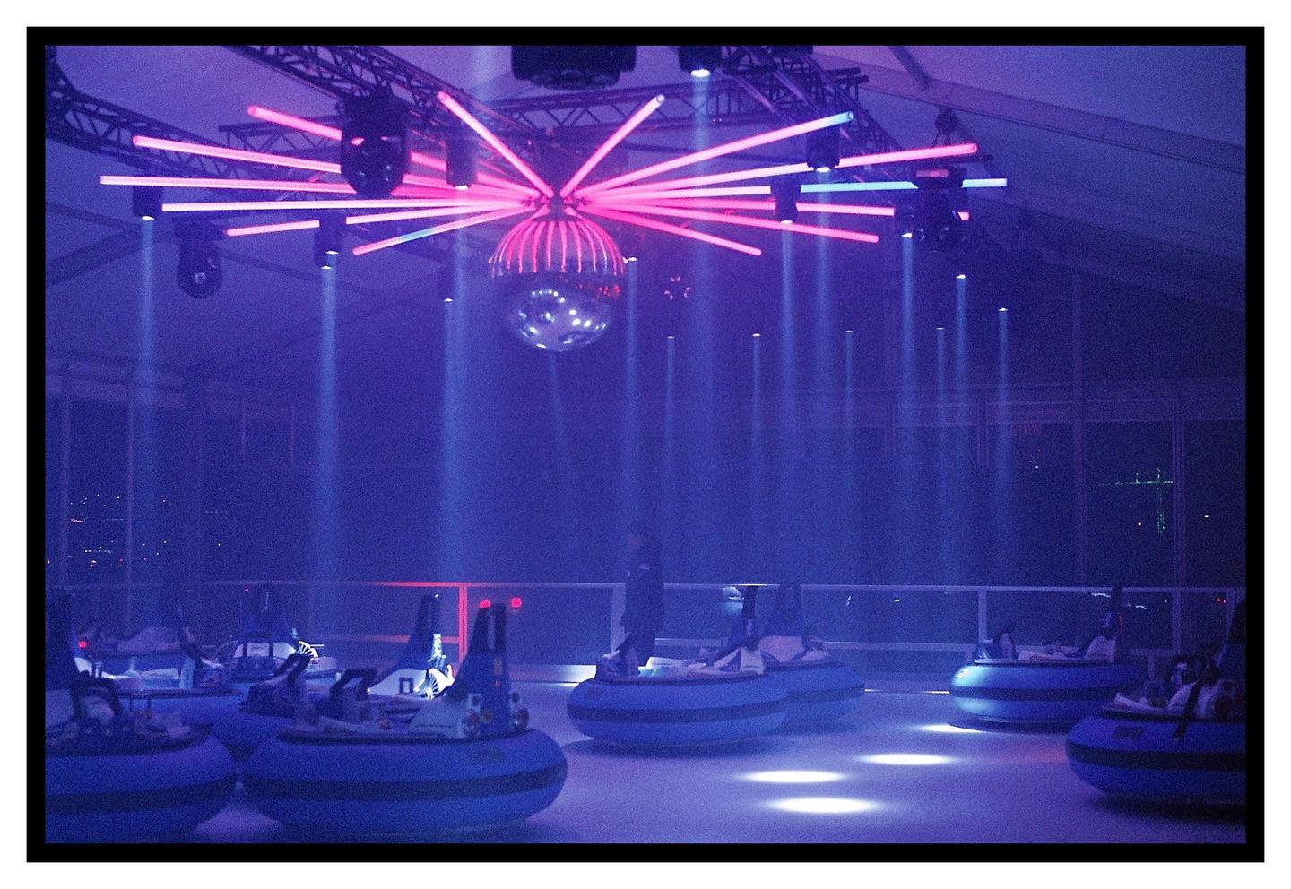 A disco ball and neon lights illuminate a bumper car rink at night.