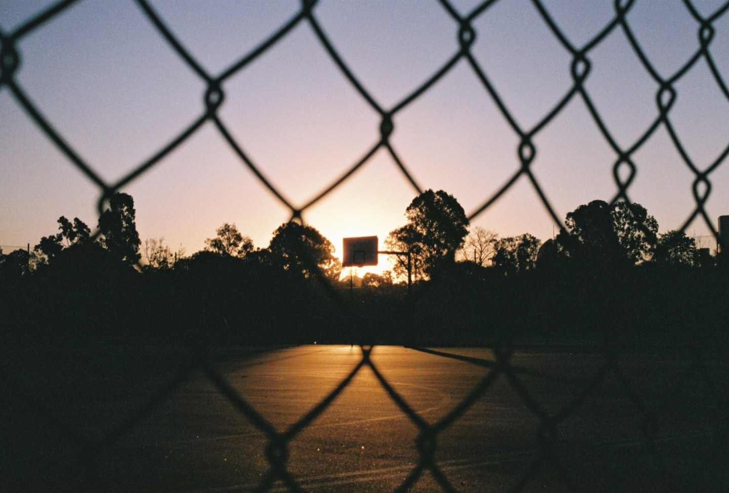 A basketball hoop is silhouetted against the sunset, viewed through a chain link fence.