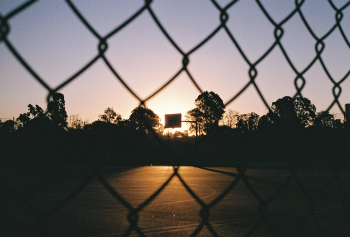 A basketball hoop is silhouetted against the sunset, viewed through a chain link fence.