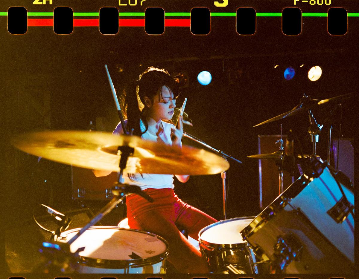A woman plays a drum kit on a dark stage.