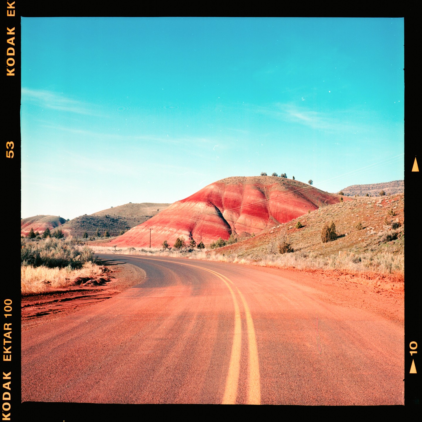 A winding road leads towards a red and orange striped hill under a clear blue sky.