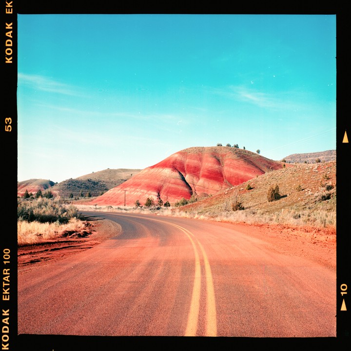 A winding road leads towards a red and orange striped hill under a clear blue sky.