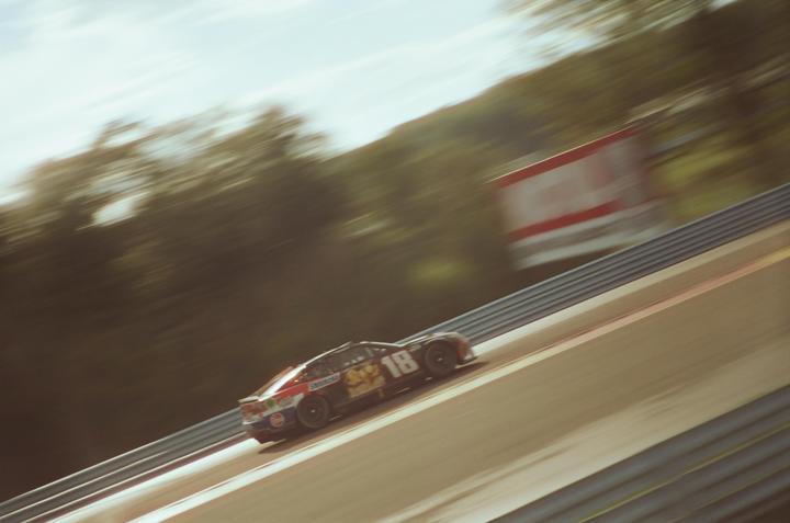 A race car with the number 18 on its side speeds along a track with blurred trees and a sign in the background.