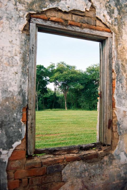 A view through a broken window frame reveals a grassy field and trees under a clear sky.