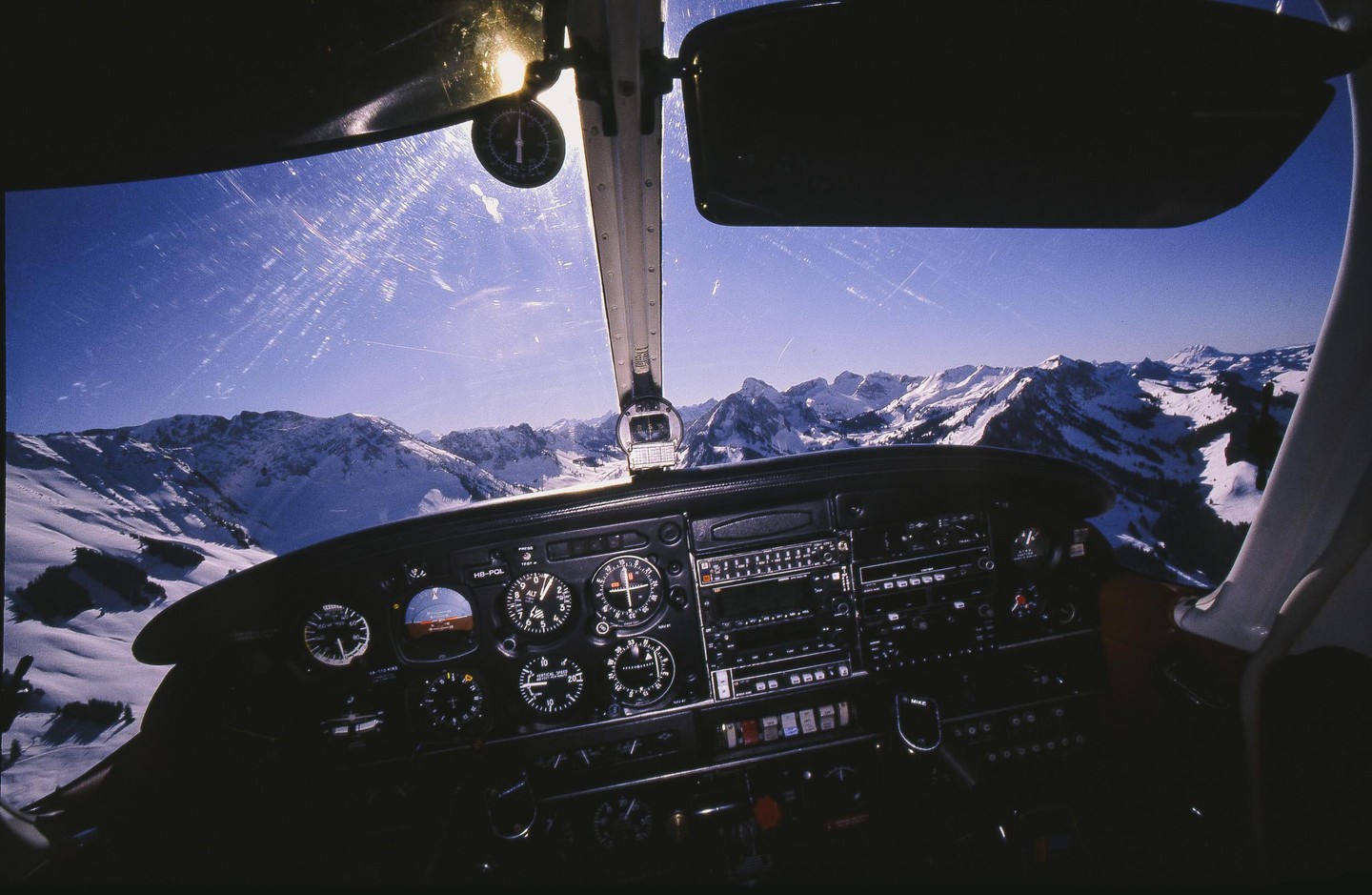 The cockpit of a small aircraft is visible with a view of snow-covered mountains through the windshield.