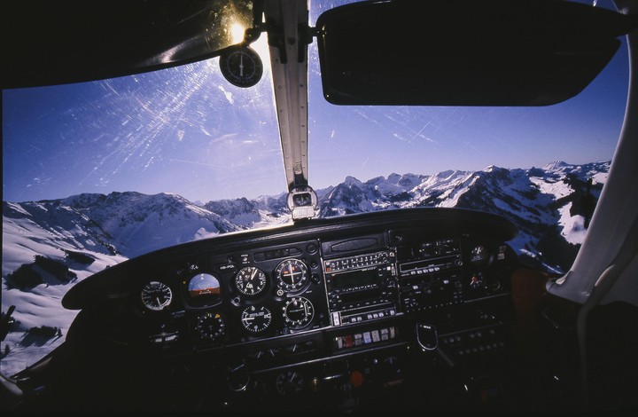 The cockpit of a small aircraft is visible with a view of snow-covered mountains through the windshield.