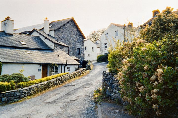 A narrow, winding road leads through a village with stone and white-washed houses under a bright sky.