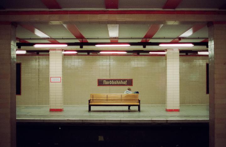 A person sits on a wooden bench in a subway station with a "Nordbahnhof" sign on the wall.