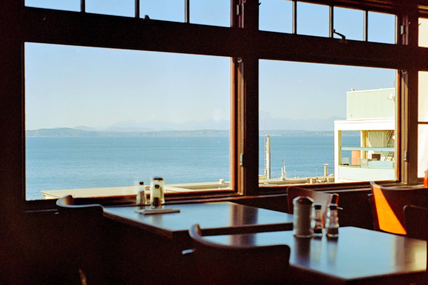 A restaurant interior with tables and chairs looks out through large windows onto a body of water with distant land and mountains.