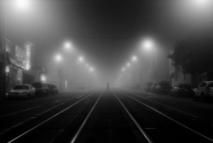 A lone man stands in the middle of a foggy street with tram tracks and parked cars on either side.