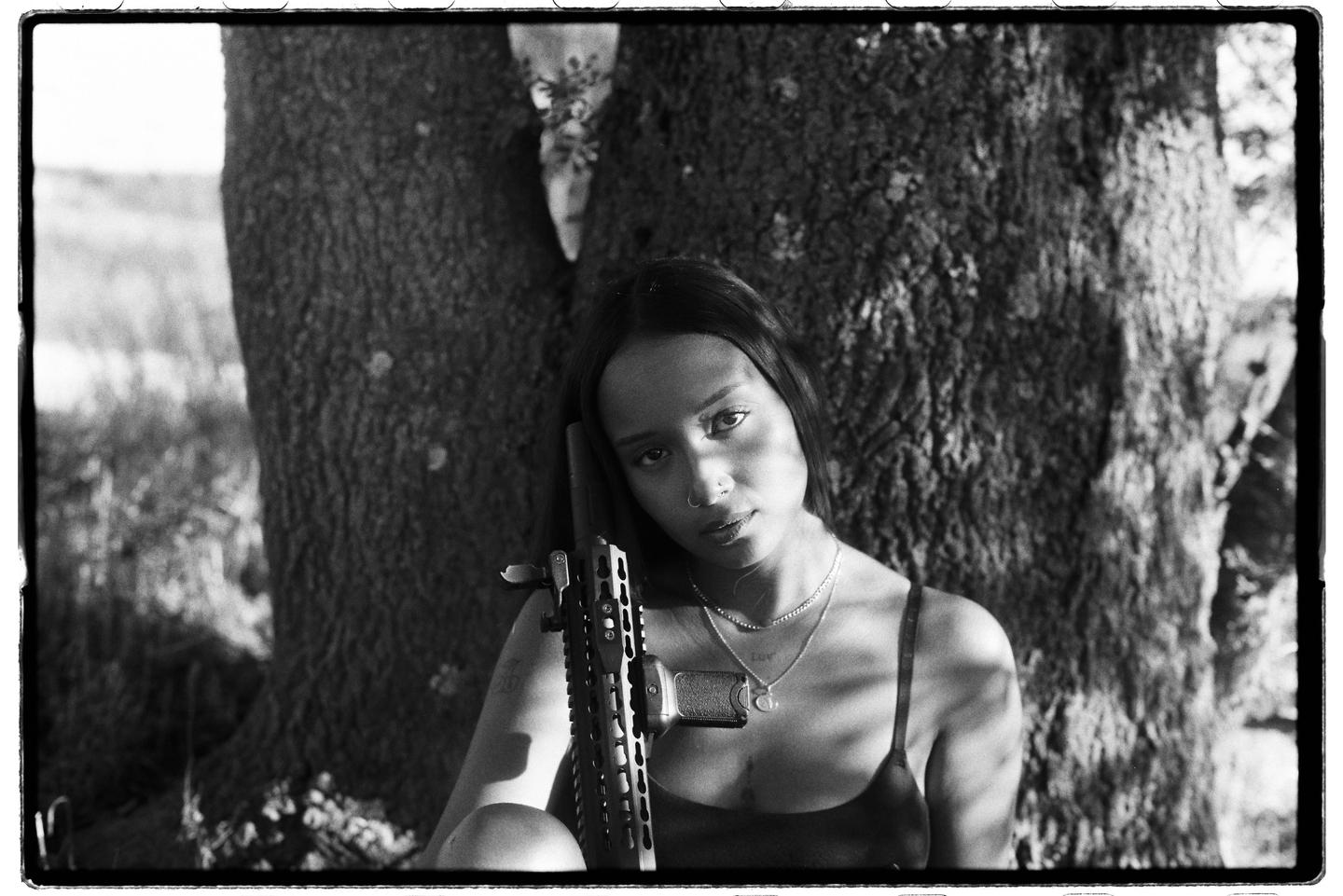 A woman with long hair and a nose ring holds a rifle while sitting in front of a tree.