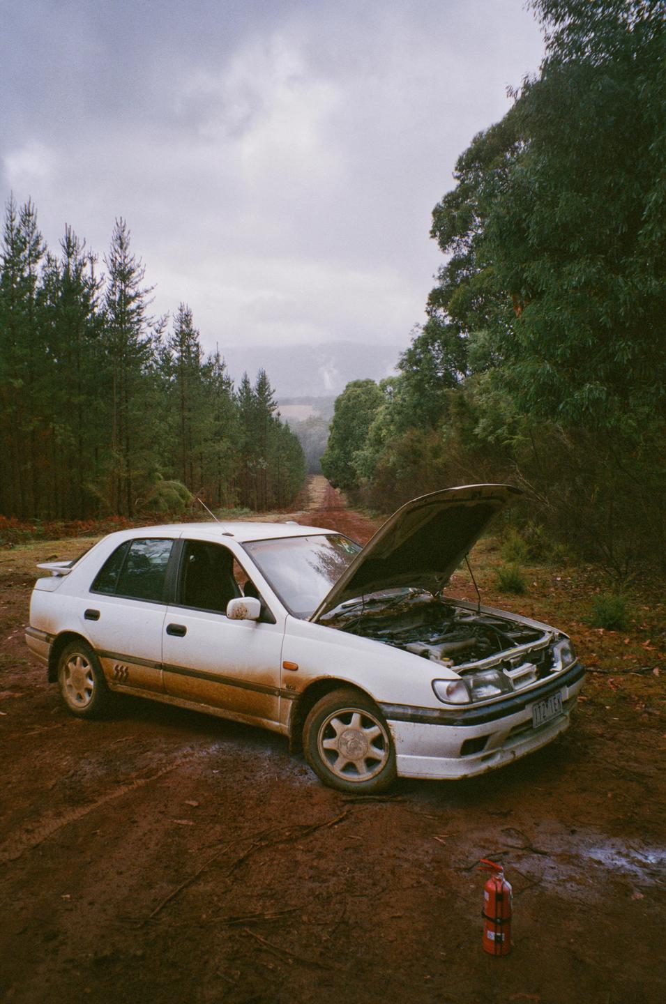 A white car with its hood open is parked on a muddy dirt road next to a forest, with a fire extinguisher on the ground in front of it.