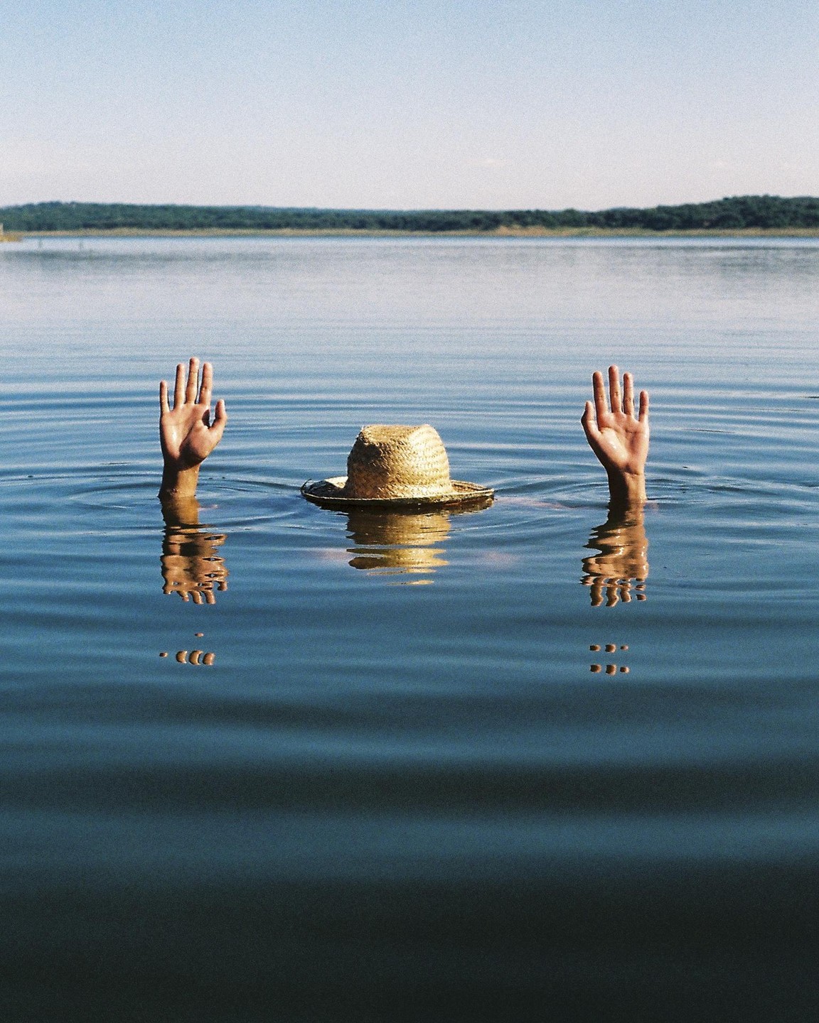Two hands and a straw hat emerge from the water of a lake with a distant treeline under a clear sky.