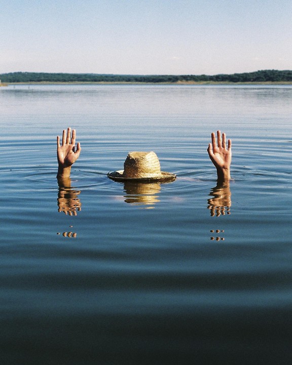 Two hands and a straw hat emerge from the water of a lake with a distant treeline under a clear sky.