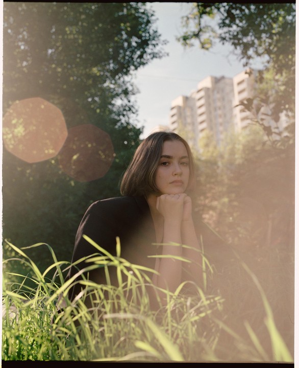 A woman with short hair rests her chin on her hands, looking directly forward, with tall grass in the foreground and apartment buildings in the background.