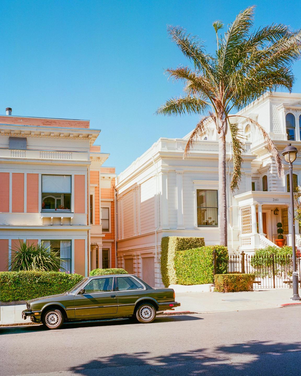 A green car is parked on a street in front of two houses and a palm tree.