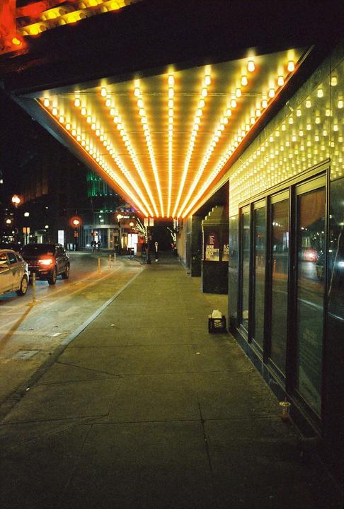 A brightly lit marquee extends over a city sidewalk at night, with cars driving on the street.