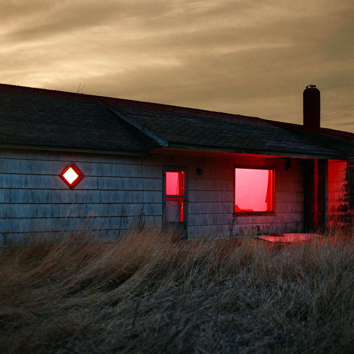 An abandoned house with glowing red windows stands in a field of tall, dry grass under a cloudy sky.