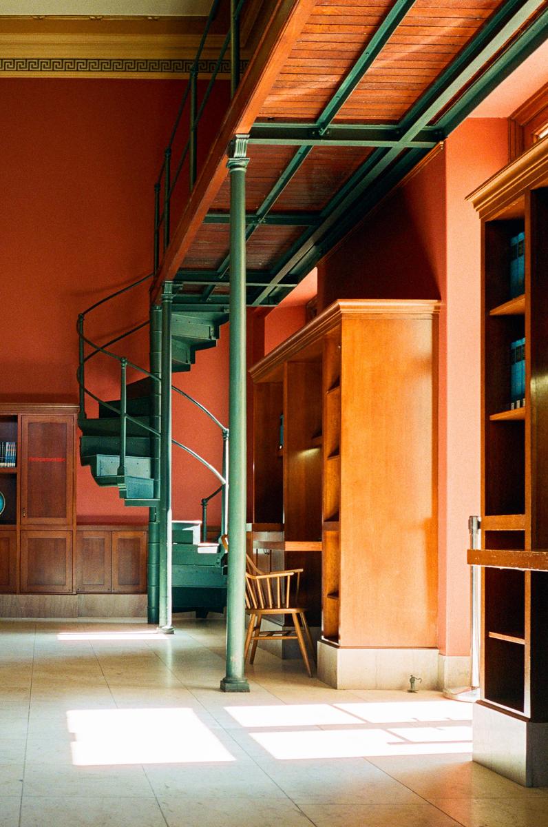 A spiral staircase leads to a second-story walkway in a room with red walls and wooden bookshelves.