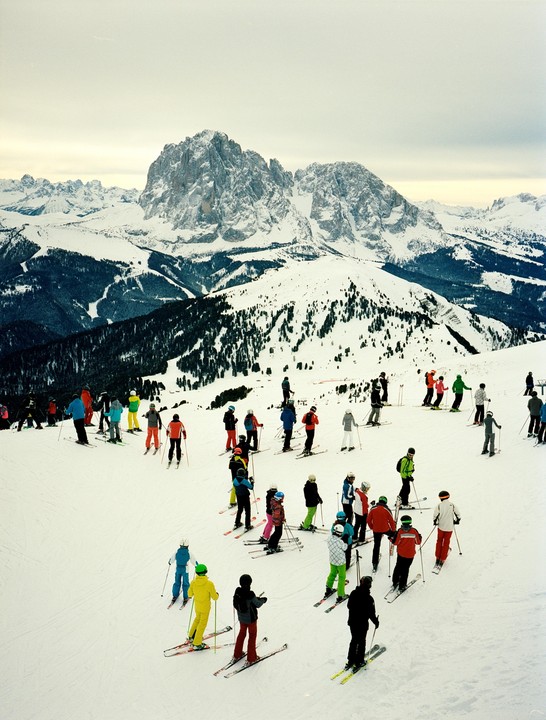 A group of skiers in colorful attire stands on a snowy mountain slope with large, rugged peaks in the background.