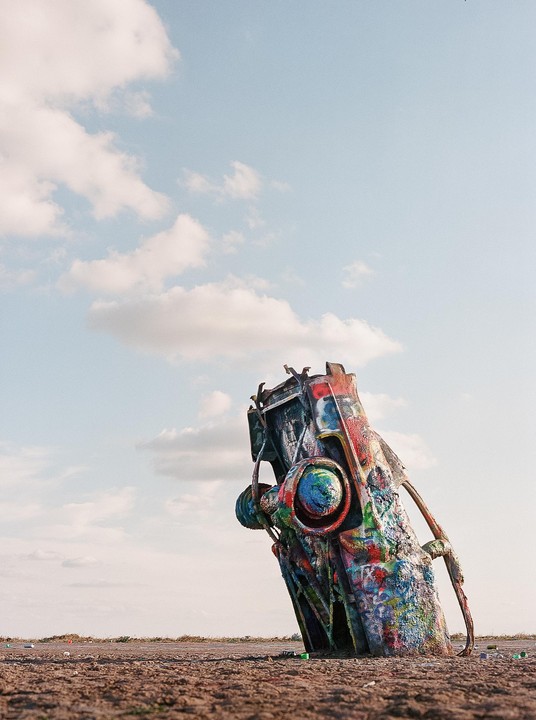 A colorful, graffiti-covered car is buried nose-down in the ground under a partly cloudy sky.